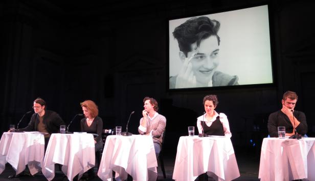 Fünf Personen sitzen an Tischen auf einer Bühne vor einem Porträtgemälde.