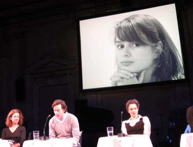 Auf einer Bühne sitzen drei Personen vor einer Leinwand mit dem Porträt einer jungen Frau.