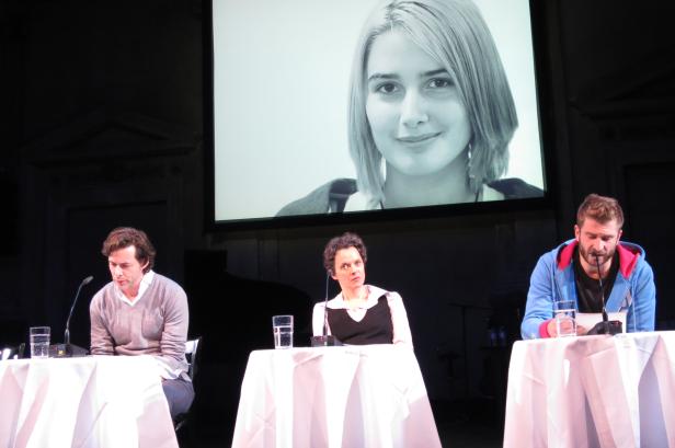 Drei Personen sitzen an Tischen vor einer Leinwand mit dem Porträt einer jungen Frau.