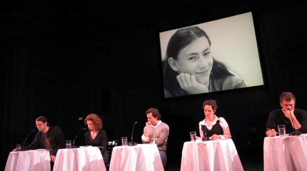 Fünf Personen sitzen an Tischen auf einer Bühne vor einem großen Bildschirm mit einem Porträt einer jungen Frau.