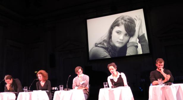 Fünf Personen sitzen an Tischen vor einer Leinwand mit dem Porträt einer jungen Frau.