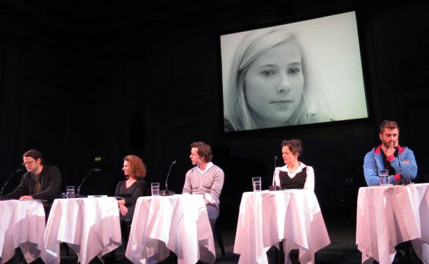 Fünf Personen sitzen an Tischen auf einer Bühne vor einem großen Bildschirm mit einem Porträt einer jungen Frau.