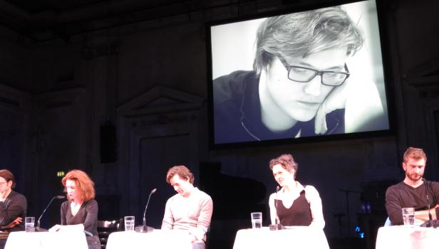 Fünf Personen sitzen an Tischen auf einer Bühne vor einer Leinwand mit einem Porträt.