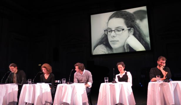 Fünf Personen sitzen an Tischen auf einer Bühne, im Hintergrund ein Porträt einer jungen Frau.