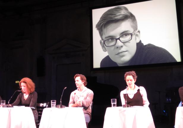 Drei Personen sitzen an Tischen vor einer Leinwand mit einem Porträt.