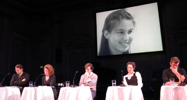 Fünf Personen sitzen an Tischen vor einer Leinwand mit einem Porträt.