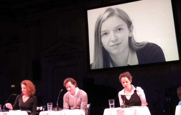 Drei Personen sitzen an einem Tisch während einer Podiumsdiskussion, im Hintergrund ein Porträt auf einer Leinwand.