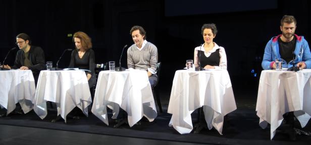 Fünf Personen sitzen an Tischen mit weißen Tischdecken auf einer Bühne.