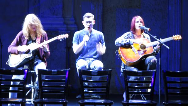 Drei Musiker sitzen auf Stühlen und spielen Gitarre und singen.
