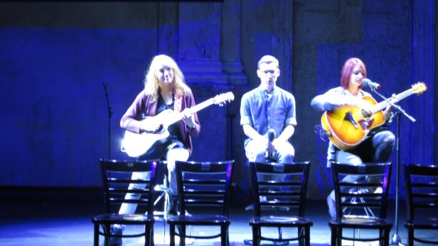 Drei Musiker sitzen auf Stühlen und spielen Gitarre auf einer Bühne.