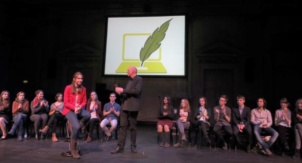Eine junge Frau sitzt auf der Bühne, während ein Mann spricht und das Publikum applaudiert.