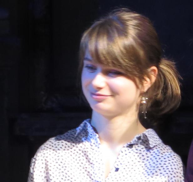 Eine junge Frau mit blonden Haaren und Ohrringen lächelt.