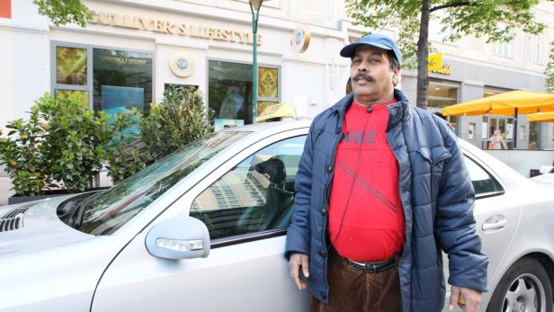 Ein Taxifahrer steht vor seinem silbernen Taxi in einer städtischen Umgebung.