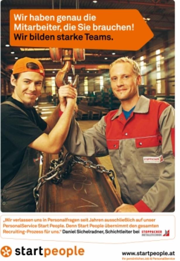 Zwei Arbeiter in Arbeitskleidung halten gemeinsam eine schwere Metallstange mit einer Kette hoch.