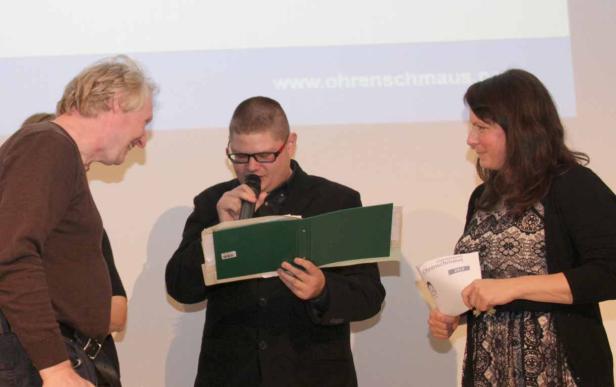 Ein Mann mit Mikrofon liest aus einem Ordner, während zwei Personen ihm aufmerksam zuhören.