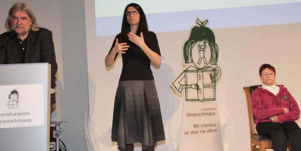 Eine Frau gebärdet auf einer Bühne, während ein Mann an einem Rednerpult steht und eine weitere Frau sitzt.