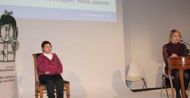 Eine Frau sitzt auf einem Stuhl neben einem Plakat, während eine andere Frau an einem Rednerpult steht.