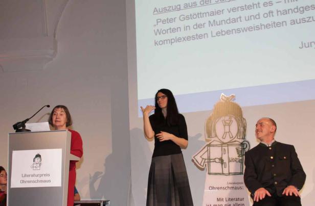 Eine Frau hält eine Rede, während eine andere in Gebärdensprache übersetzt; ein Mann im Trachtenjanker sitzt daneben.