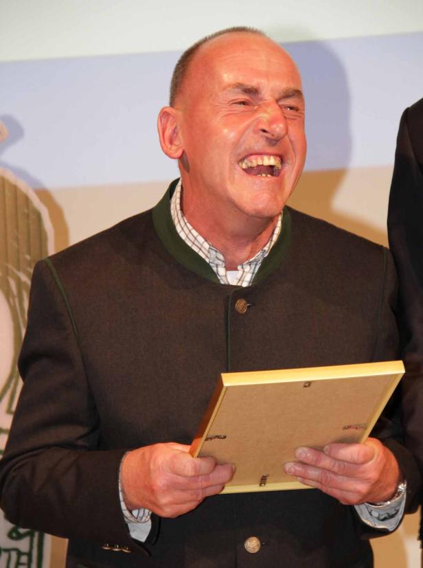 Ein Mann in Trachtenjacke hält lachend einen Bilderrahmen in den Händen.