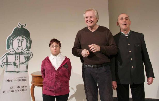 Drei Personen stehen neben einer Zeichnung des Literaturpreises Ohrenschmaus.