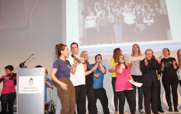 Eine Gruppe von Menschen tritt auf einer Bühne auf, einige singen ins Mikrofon, während andere applaudieren.