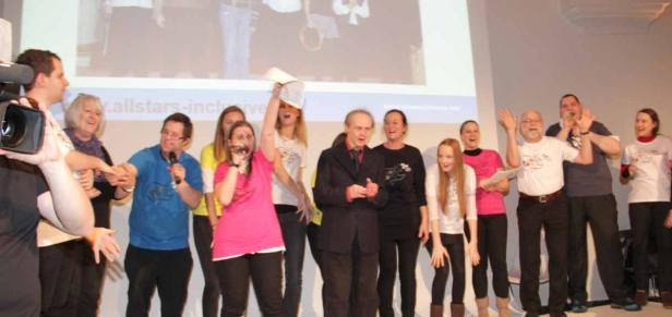 Eine Gruppe von Menschen steht auf einer Bühne und feiert mit Mikrofonen und erhobenen Händen.