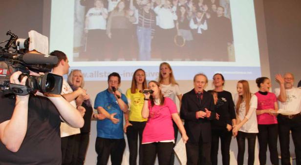 Eine Gruppe von Menschen steht auf einer Bühne und singt in Mikrofone.