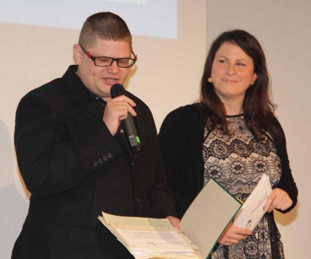 Ein Mann spricht am Mikrofon, während eine Frau mit Unterlagen daneben steht.