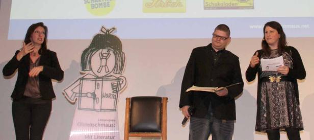 Drei Personen stehen auf einer Bühne neben einer Cartoon-Figur für den Literaturpreis „Ohrenschmaus“.