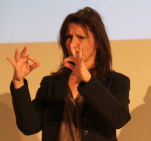 Eine Frau gestikuliert mit ihren Händen vor einem hellen Hintergrund.