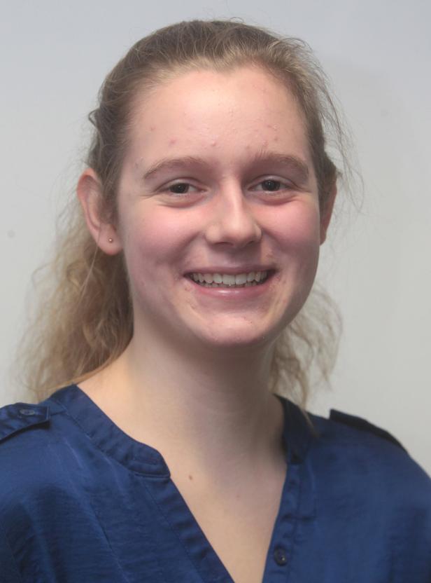 Eine lächelnde junge Frau mit blonden Haaren und einem blauen Hemd vor einem hellen Hintergrund.