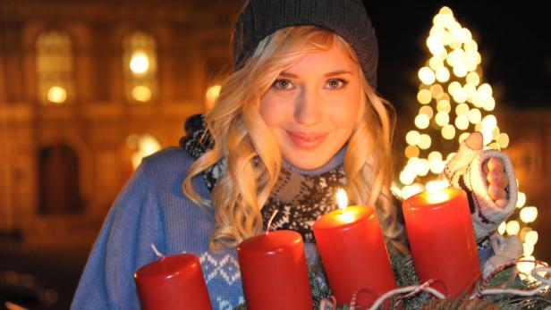 Eine blonde Frau mit Adventskranz und brennenden Kerzen vor einem erleuchteten Gebäude.