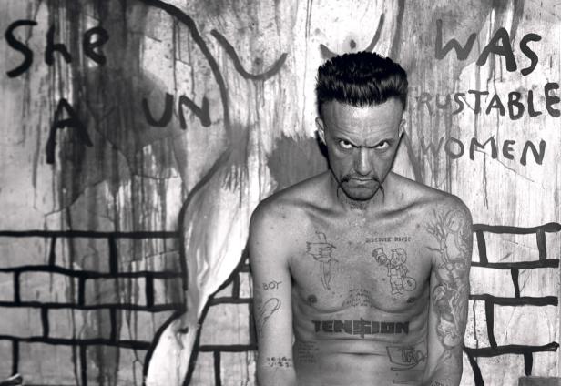 Ein Mann mit Tätowierungen steht vor einer Wand mit Graffiti.
