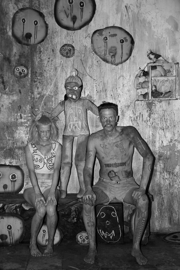 Schwarzweißaufnahme einer Frau, eines Mannes und einer gruseligen Puppe vor einer dekorierten Wand.