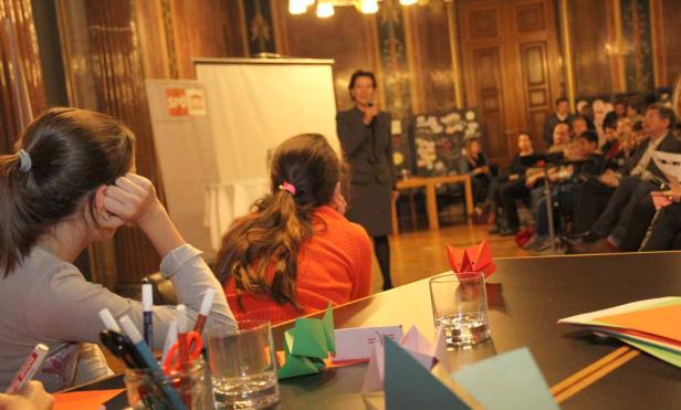 Eine Frau hält eine Rede vor einem Publikum, während Kinder am Tisch sitzen und basteln.