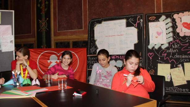 Eine Gruppe von Mädchen und eine Frau sitzen an einem Tisch und basteln vor einer Tafel.