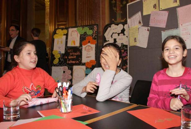 Drei Mädchen sitzen an einem Tisch und basteln vor einer Tafel mit Kinderzeichnungen.