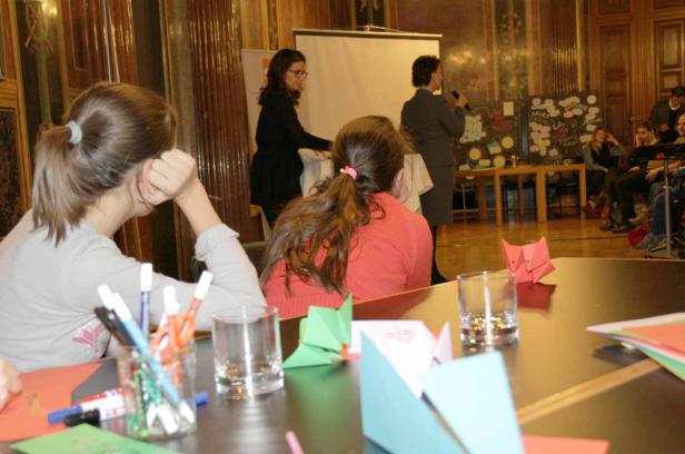 Zuschauer sitzen an Tischen mit Origami, während zwei Frauen auf einer Bühne sprechen.