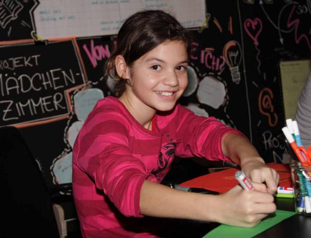 Ein junges Mädchen zeichnet an einem Tisch vor einer Tafel mit Kritzeleien.