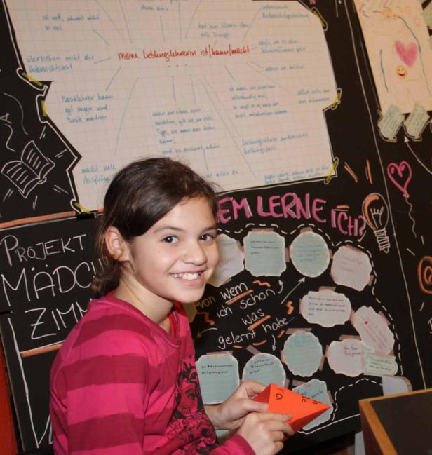 Ein lächelndes Mädchen steht vor einer Tafel mit dem Titel „Projekt Mädchenzimmer“.