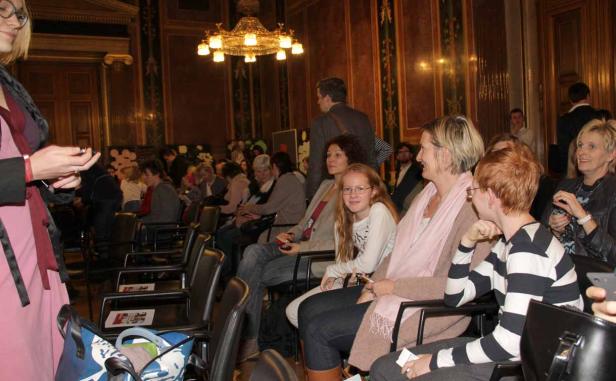 Zuschauer sitzen in einem eleganten Saal unter einem Kronleuchter.