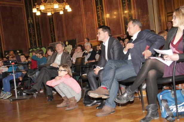 Zuschauer, darunter Kinder und Erwachsene, sitzen in einem Saal unter einem Kronleuchter.