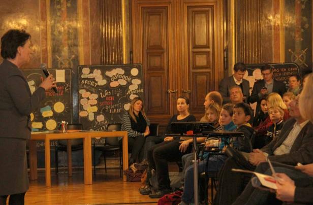 Eine Frau spricht vor einem Publikum mit Tafeln im Hintergrund.