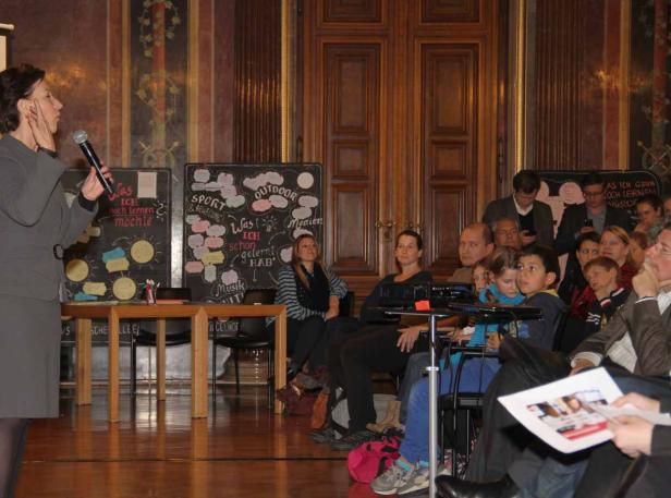 Eine Frau spricht vor einem Publikum mit Tafeln im Hintergrund.