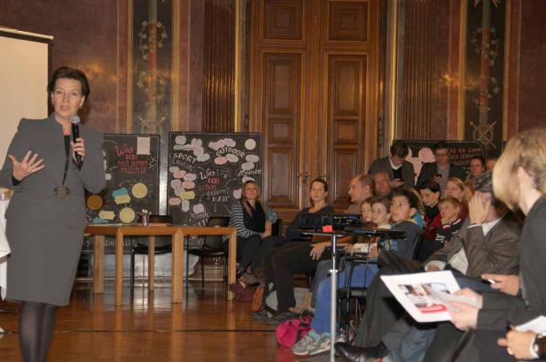 Eine Frau hält ein Mikrofon und spricht vor einem Publikum in einem Raum mit Tafeln.