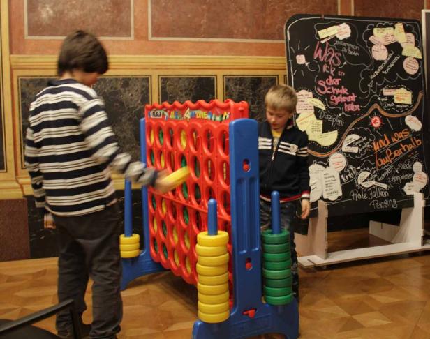 Zwei Jungen spielen „Vier gewinnt“ vor einer Tafel mit Notizen.