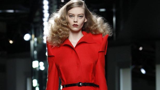 Eine Frau mit blond gelocktem Haar trägt einen roten Mantel mit einem schwarzen Gürtel auf einem Laufsteg.