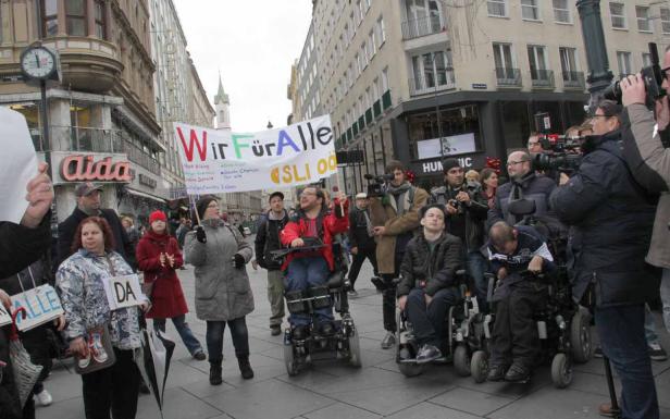 Eine Demonstration mit Menschen mit Behinderungen und einem Banner „Wir Für Alle“ in einer Stadt.