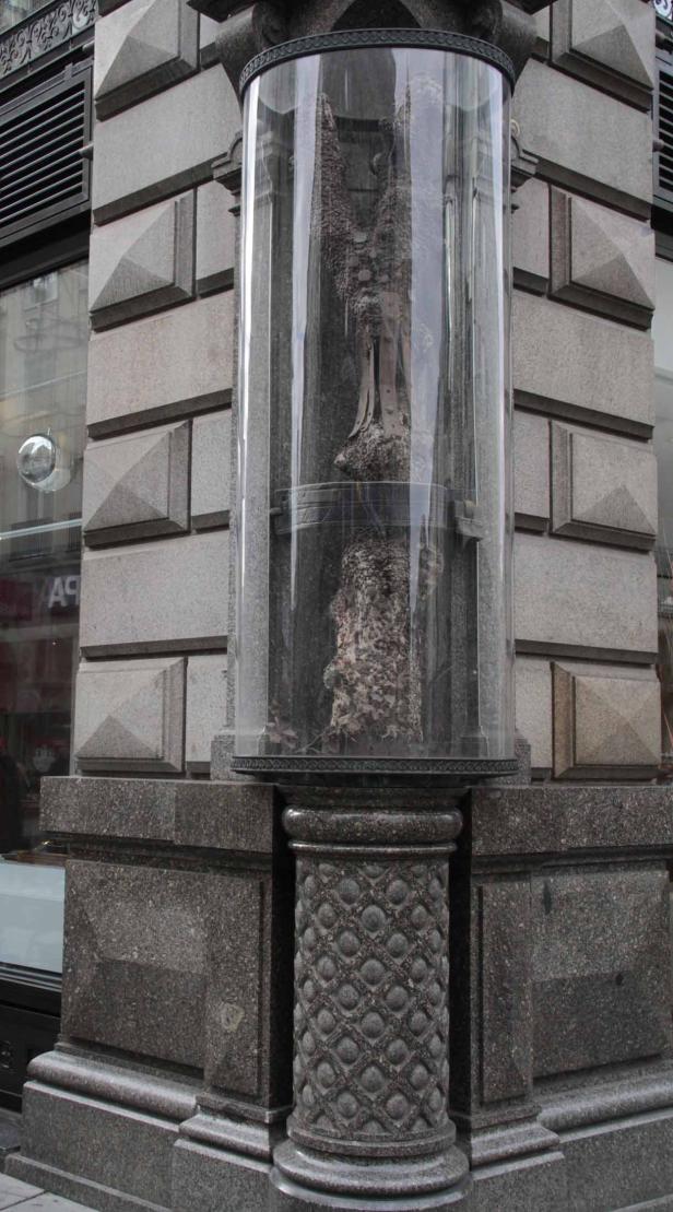 Ein Baumstumpf befindet sich in einem zylindrischen Glaskasten vor einem Gebäude.