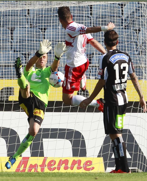 Ein Fußballtorwart versucht, einen Schuss abzuwehren, während zwei andere Spieler zusehen.